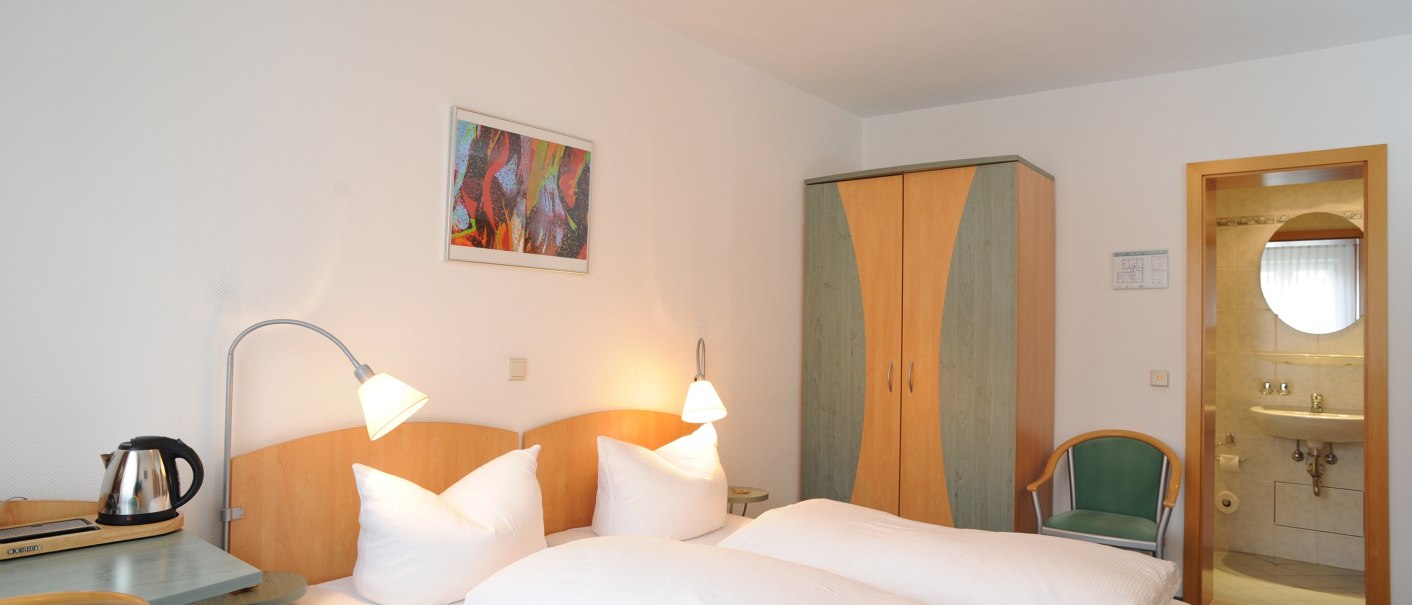 Ein gemütliches Hotelzimmer mit Doppelbett, Kleiderschrank und einem kleinen Tisch mit Wasserkocher. Ein Badezimmer ist im Hintergrund sichtbar., © TOMAS