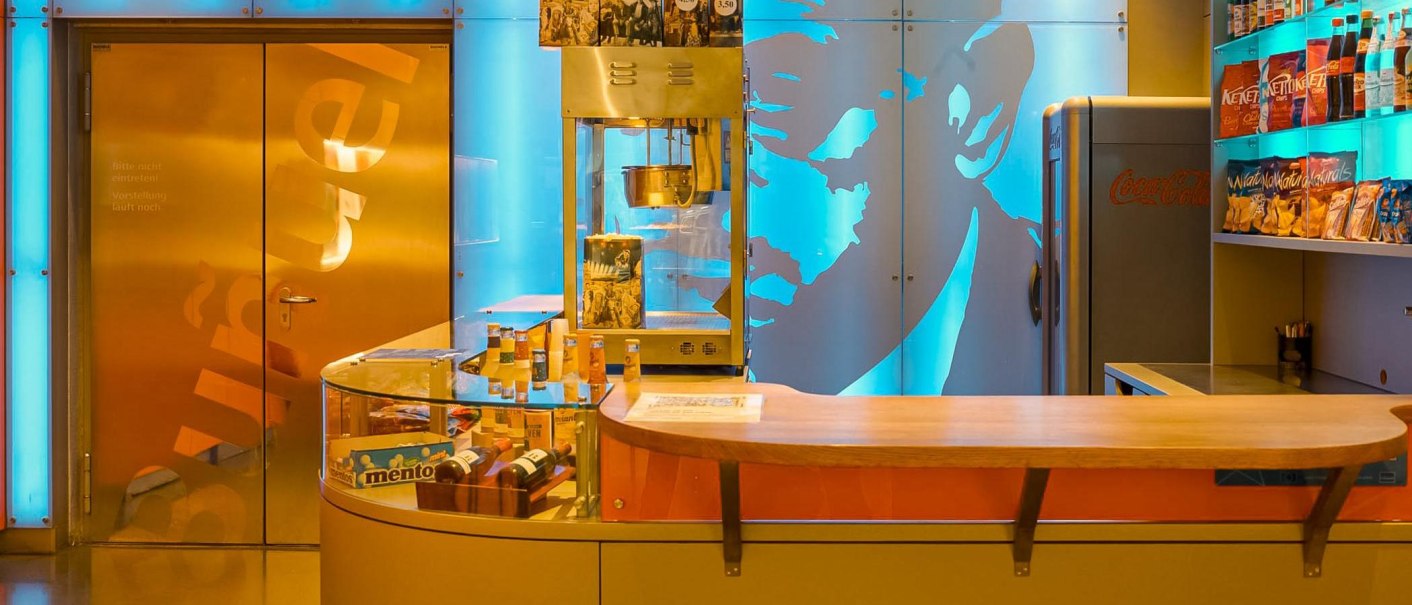 Ein Kinokiosk mit Popcornmaschine, Snacks und Getränken. Die Wände sind in Blau und Orange beleuchtet. Ein Kühlschrank steht rechts., © Delphi Arthaus, Stuttgart