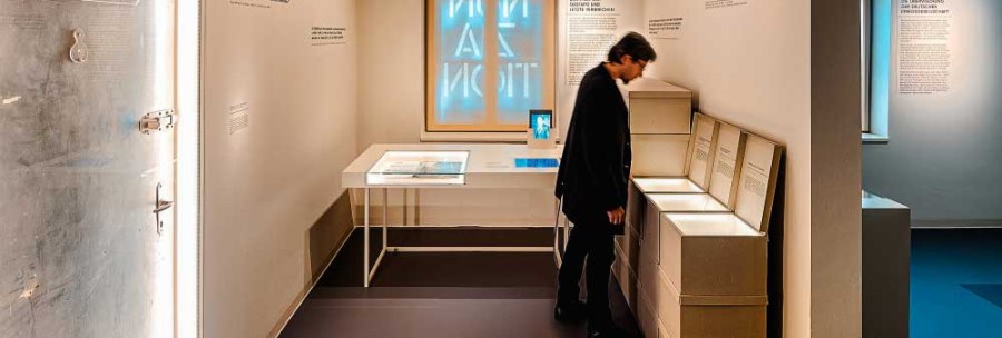A museum room with a person looking at exhibits. There are texts and an illuminated window on the wall. On the left a door with the inscription '01 Lager'., © Haus der Geschichte Baden-Württemberg