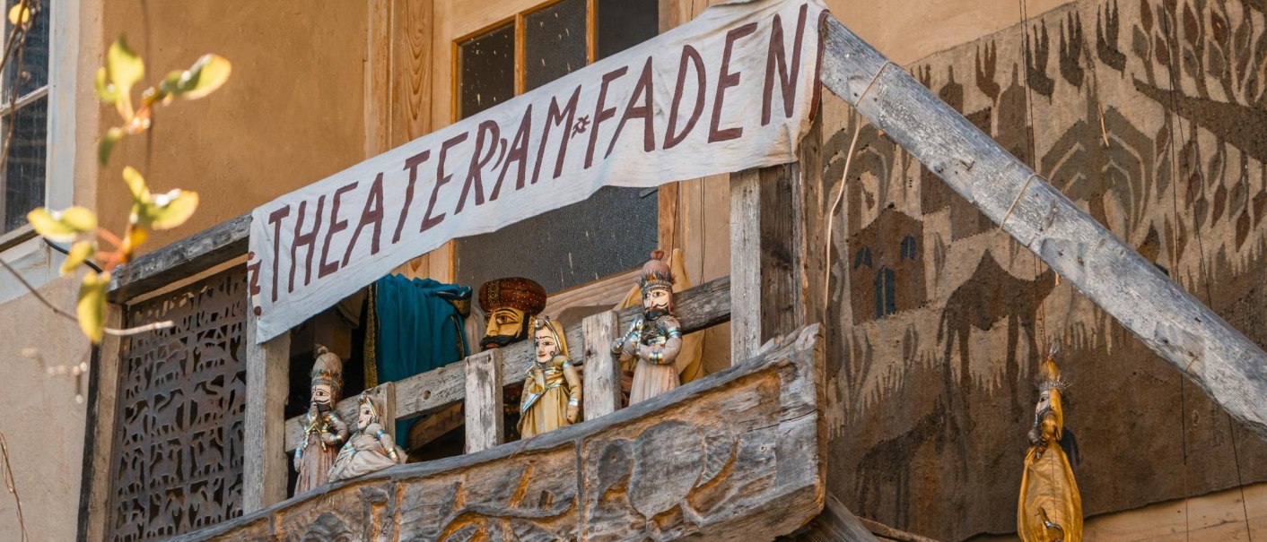 Ein Holzbalkon mit Puppen und einem handgemalten Banner 'Theater am Faden'. Die Puppen sind traditionell gekleidet., © SMG, Sarah Schmid Ein Holzbalkon mit Puppen und einem handgemalten Banner 'Theater am Faden'. Die Puppen sind traditionell gekleidet., © SMG, Sarah Schmid
