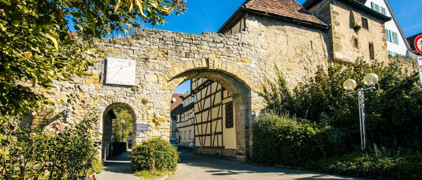 An old town gate in Marbach am Neckar, flanked by half-timbered houses and surrounded by green vegetation under a blue sky., © Stuttgart-Marketing GmbH, Sarah Schmid An old town gate in Marbach am Neckar, flanked by half-timbered houses and surrounded by green vegetation under a blue sky., © Stuttgart-Marketing GmbH, Sarah Schmid