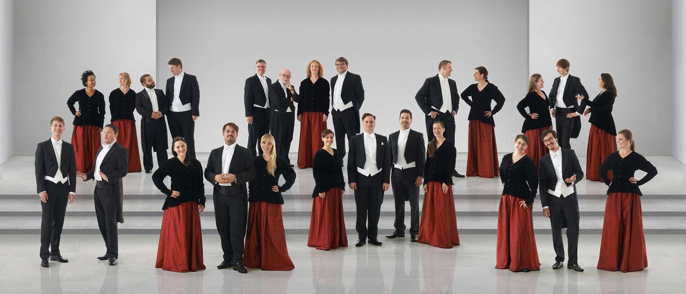 Der Kammerchor Stuttgart posiert in formeller Kleidung auf einer Bühne. Die Frauen tragen rote Röcke und schwarze Oberteile, die Männer Frack., © Jens Meisert Der Kammerchor Stuttgart posiert in formeller Kleidung auf einer Bühne. Die Frauen tragen rote Röcke und schwarze Oberteile, die Männer Frack., © Jens Meisert