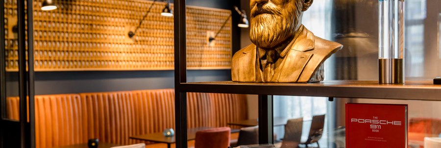 Büste eines Mannes mit Bart, ein Modellauto und ein Porsche-Buch in einem stilvoll eingerichteten Raum mit Ledersitzbänken., © TOMAS Büste eines Mannes mit Bart, ein Modellauto und ein Porsche-Buch in einem stilvoll eingerichteten Raum mit Ledersitzbänken., © TOMAS