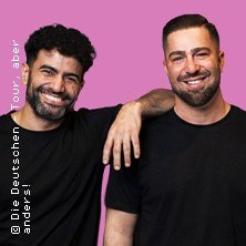 Two men in black T-shirts smile in front of a pink background. One puts his arm around the other. Text: "The Germans tour, but differently!", &copy; links im Bild
