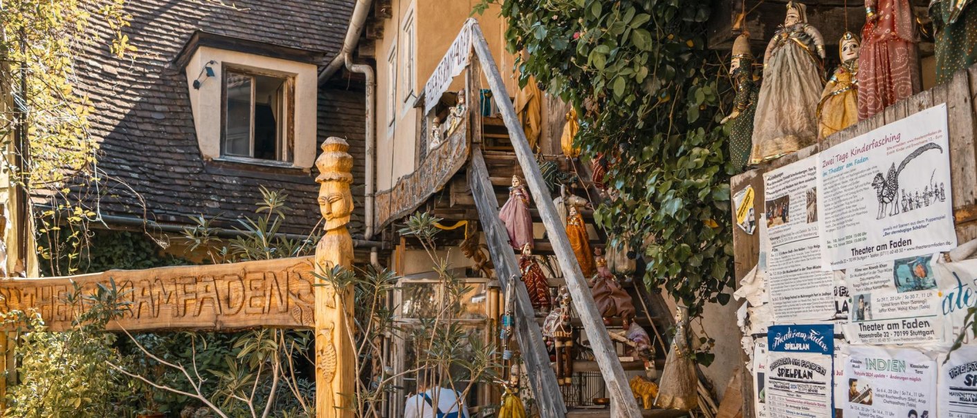 Eingang des Theater am Faden mit geschnitztem Holzschild, Puppen an einer Treppe und Plakaten an der Wand. Pflanzen umrahmen die Szene., © SMG, Sarah Schmid