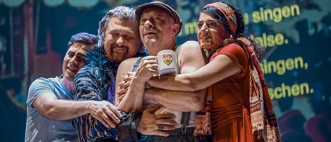 Vier Personen umarmen sich herzlich auf einer Bühne. Eine Person hält einen Becher mit einem Logo. Im Hintergrund sind verschwommene Texte projiziert., © Altes Schauspielhaus, Stuttgart