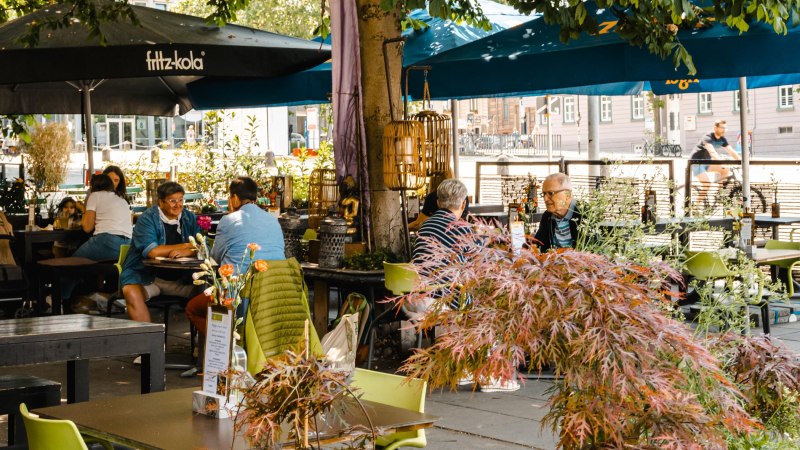 Ein belebtes Caf&eacute; im Freien mit Menschen, die an Tischen unter Sonnenschirmen sitzen. Pflanzen und Dekorationen schaffen eine gem&uuml;tliche Atmosph&auml;re., &copy; SMG Stuttgart Marketing GmbH - Sarah Schmid