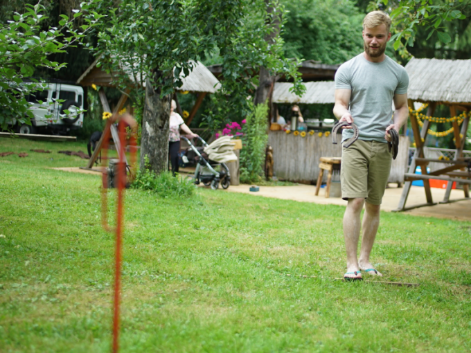 Ein Mann in Shorts und T-Shirt wirft Hufeisen auf einer Wiese. Im Hintergrund sind Bäume und eine Hütte zu sehen., © Cool-Tours StattReisen Ein Mann in Shorts und T-Shirt wirft Hufeisen auf einer Wiese. Im Hintergrund sind Bäume und eine Hütte zu sehen., © Cool-Tours StattReisen