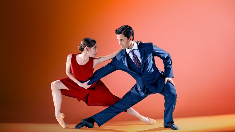 Ein Tänzerpaar in eleganter Pose, sie im roten Kleid, er im Anzug, vor einem orangefarbenen Hintergrund. Beide blicken sich intensiv an., © Württembergische Staatstheater Stuttgart Ein Tänzerpaar in eleganter Pose, sie im roten Kleid, er im Anzug, vor einem orangefarbenen Hintergrund. Beide blicken sich intensiv an., © Württembergische Staatstheater Stuttgart