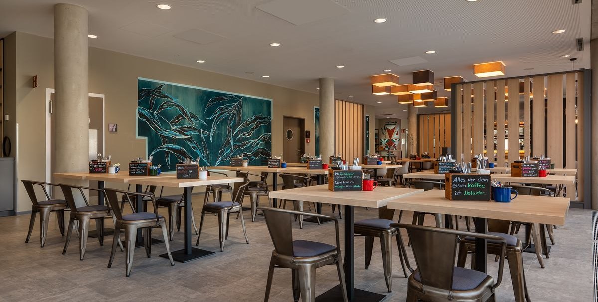 Moderner Frühstücksraum mit Holztischen, Metallstühlen und einem großen Wandbild. Die Beleuchtung ist warm und einladend., © Novum Hospitality