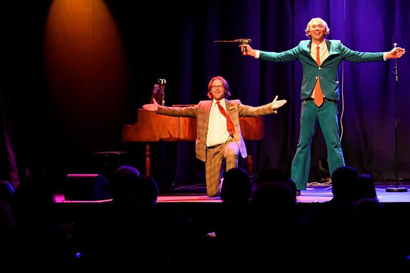Zwei M&auml;nner in bunten Anz&uuml;gen performen auf einer B&uuml;hne. Einer kniet vor einem Klavier, der andere steht mit ausgebreiteten Armen. Publikum im Vordergrund., &copy; Theaterhaus Stuttgart e.V.