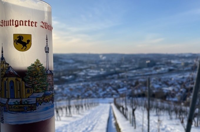Ein Glas Glühwein mit Stuttgarter Motiv steht vor einer verschneiten Weinberglandschaft. Im Hintergrund ist die Stadt Stuttgart zu sehen., © Weingenuss Stuttgart