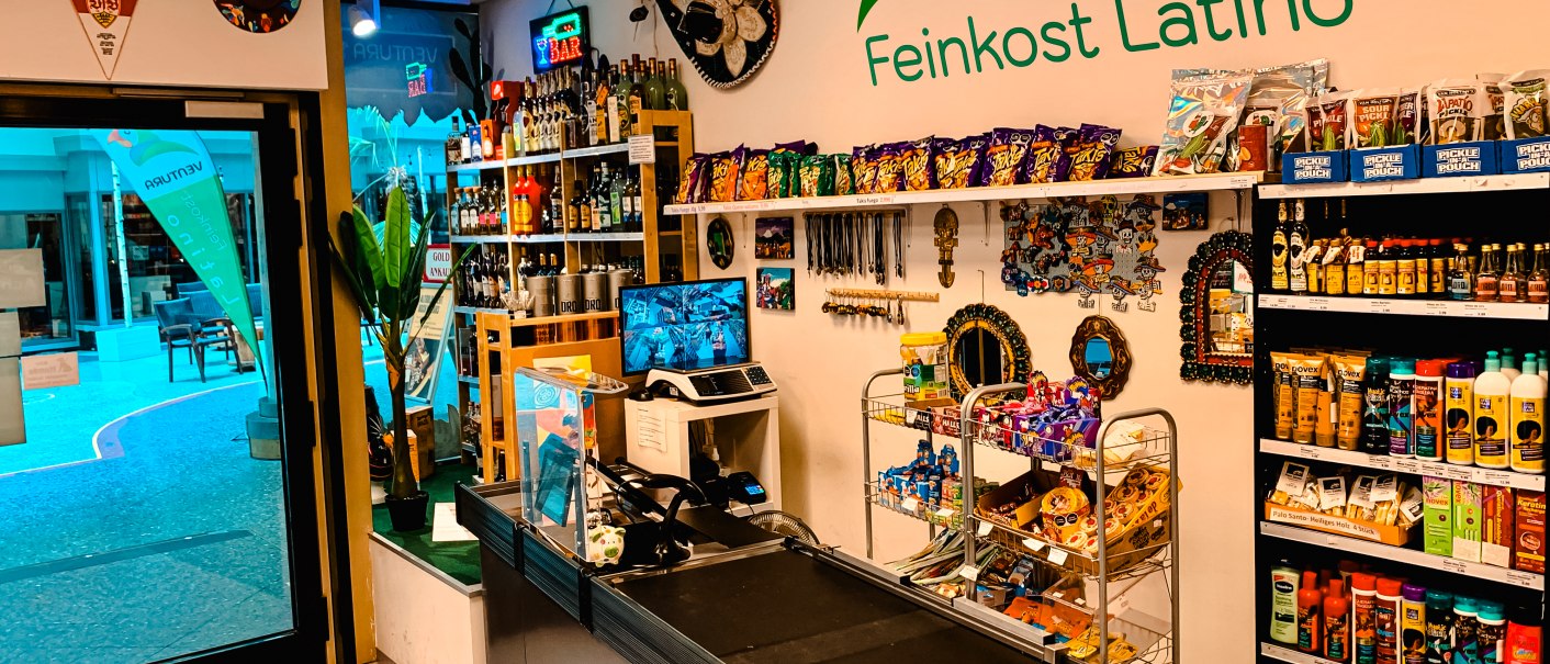Ein Feinkostladen mit lateinamerikanischen Produkten, bunten Dekorationen und einer Kasse. Regale sind mit Snacks und Getränken gefüllt., © Feinkost Latino Ventura