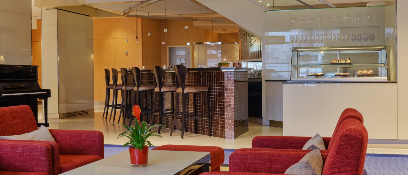 Modern hotel lobby with red armchairs, a table with a plant, a cake display case and a bar with high stools., © Steigenberger Graf Zeppelin