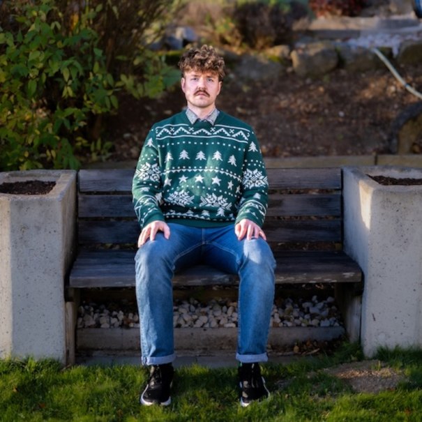 A man with curly hair is sitting on a bench outside. He is wearing a green Christmas sweater with white patterns and blue jeans., &copy; Renitenztheater Stuttgart e.V.