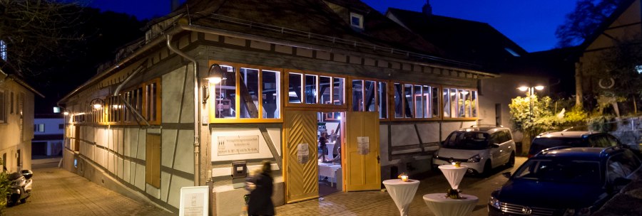 Half-timbered building at night, illuminated windows, open doors. In front of it are bar tables with white tablecloths. People and cars can be seen., © Steilwerk Rohracker Half-timbered building at night, illuminated windows, open doors. In front of it are bar tables with white tablecloths. People and cars can be seen., © Steilwerk Rohracker