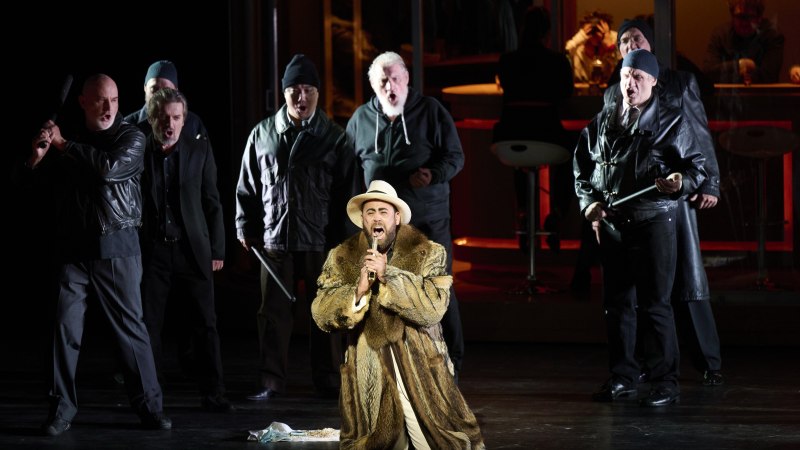 Ein Mann in Pelzmantel und Hut kniet auf der Bühne, umgeben von Männern in dunkler Kleidung. Dramatische Opernszene mit intensiver Beleuchtung., © Martin Sigmund Ein Mann in Pelzmantel und Hut kniet auf der Bühne, umgeben von Männern in dunkler Kleidung. Dramatische Opernszene mit intensiver Beleuchtung., © Martin Sigmund