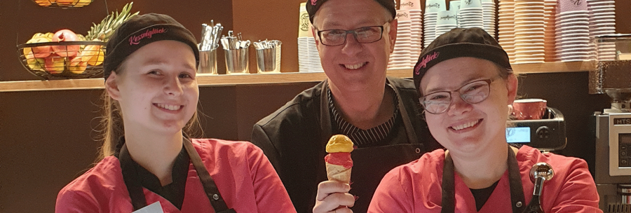 Drei Personen in einer Eisdiele lächeln in die Kamera. Eine Person hält ein Eis, im Hintergrund sind bunte Becher und Obst zu sehen., © kesselglueck.de