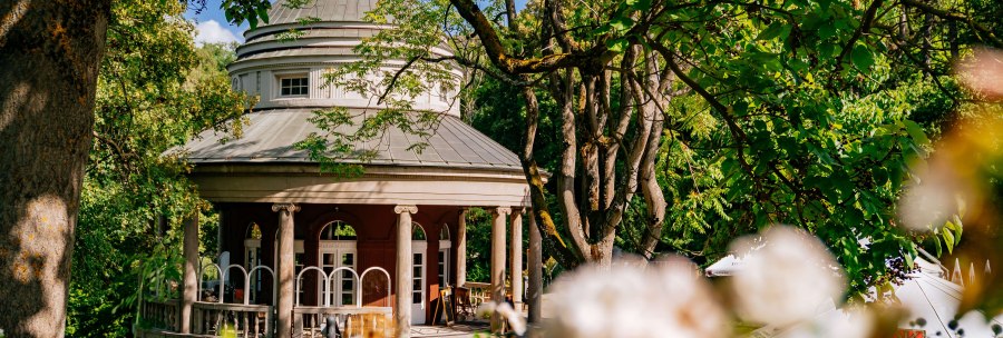 A round teahouse with pillars in Weissenburg Park, surrounded by green trees and blooming flowers in the foreground., © Thomas Niedermüller