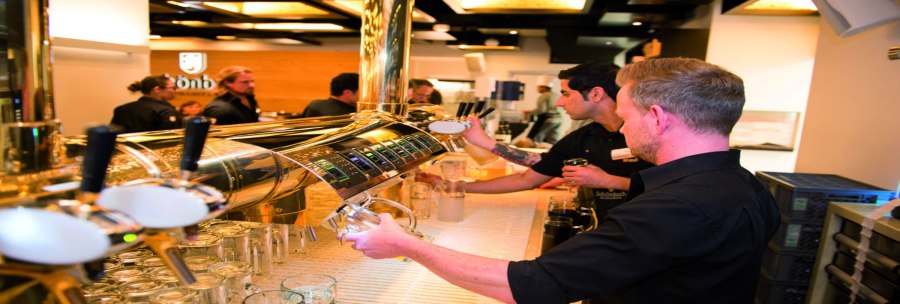 Barkeeper zapfen Bier an einer glänzenden Theke. Im Hintergrund sind weitere Personen und eine stilvolle Decke zu sehen., © Schönbuchbrauerei, Sebastian Berger