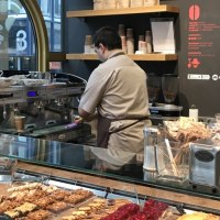 A barista in a café prepares coffee. In front of him are pastries, a coffee machine and packets of tea., © Old Bridge, Stuttgart A barista in a café prepares coffee. In front of him are pastries, a coffee machine and packets of tea., © Old Bridge, Stuttgart