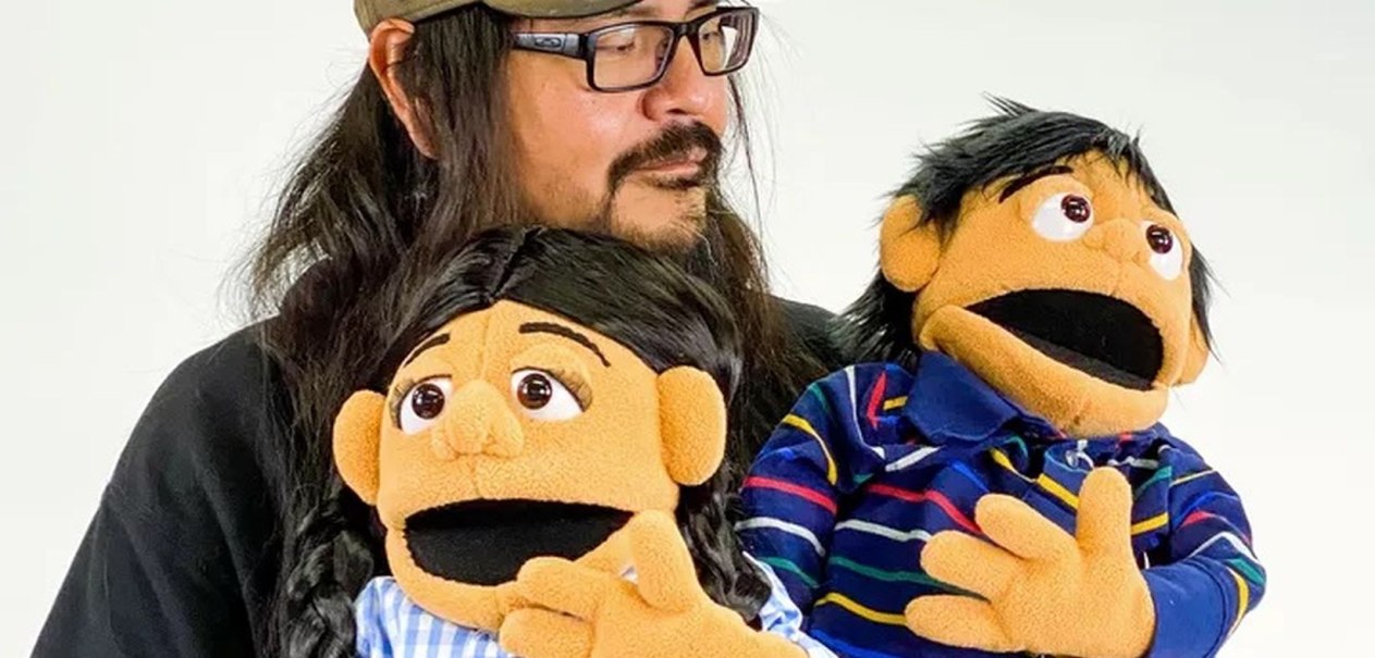 Ein Mann mit Brille und Bart hält zwei Puppen, eine in einem blauen Hemd und eine in einem karierten Hemd, vor einem weißen Hintergrund., © Pete Sands