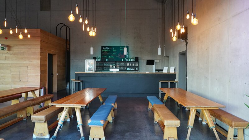 Ein modernes Café mit langen Holztischen, Hängelampen und einer Bar im Hintergrund. Die Einrichtung ist minimalistisch und einladend., © Württembergische Staatstheater Stuttgart Ein modernes Café mit langen Holztischen, Hängelampen und einer Bar im Hintergrund. Die Einrichtung ist minimalistisch und einladend., © Württembergische Staatstheater Stuttgart