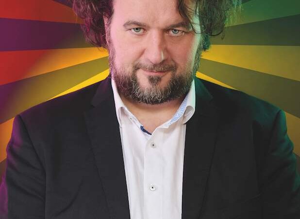 Ein Mann mit lockigem Haar und Bart tr&auml;gt einen Anzug. Der Hintergrund ist bunt und strahlenf&ouml;rmig gestaltet., &copy; Rosenau Kultur e.V.