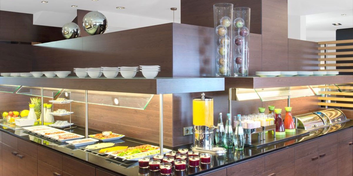 Modernes Frühstücksbuffet mit Obst, Getränken und Geschirr. Dunkle Holzmöbel und Glasdekorationen schaffen eine elegante Atmosphäre., © NH Stuttgart Sindelfingen Modernes Frühstücksbuffet mit Obst, Getränken und Geschirr. Dunkle Holzmöbel und Glasdekorationen schaffen eine elegante Atmosphäre., © NH Stuttgart Sindelfingen