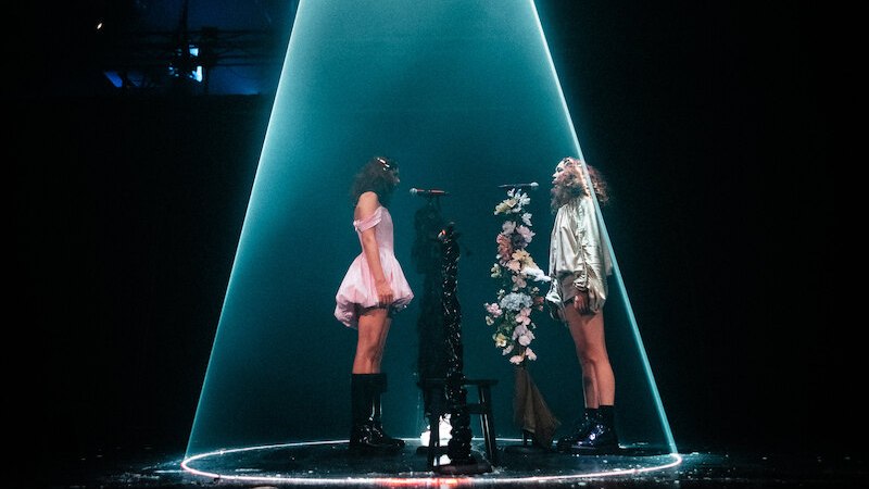 Zwei Performerinnen stehen sich auf einer Bühne gegenüber, beleuchtet von einem Lichtkegel. Zwischen ihnen ein mit Blumen geschmückter Mikrofonständer., © Theater Rampe Zwei Performerinnen stehen sich auf einer Bühne gegenüber, beleuchtet von einem Lichtkegel. Zwischen ihnen ein mit Blumen geschmückter Mikrofonständer., © Theater Rampe