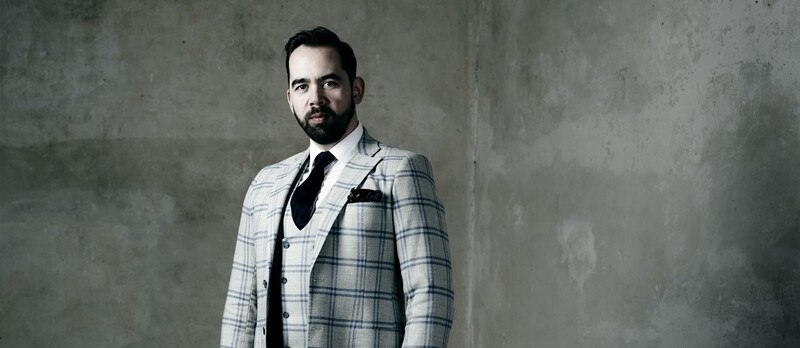 Ein Mann in einem karierten Anzug mit Krawatte steht vor einer grauen, strukturierten Wand., © Württembergische Staatstheater Stuttgart Ein Mann in einem karierten Anzug mit Krawatte steht vor einer grauen, strukturierten Wand., © Württembergische Staatstheater Stuttgart