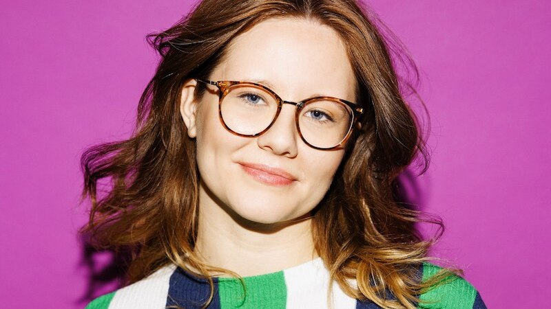 Eine Frau mit braunen Haaren und Brille l&auml;chelt vor einem lila Hintergrund. Sie tr&auml;gt einen gestreiften Pullover in Gr&uuml;n, Blau und Wei&szlig;., &copy; Theaterhaus Stuttgart e.V.