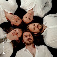 F&uuml;nf Personen in wei&szlig;en Hemden liegen im Kreis auf dem Boden und blicken nach oben. Sie tragen alle lange Haare und haben ernste Gesichtsausdr&uuml;cke., &copy; links im Bild