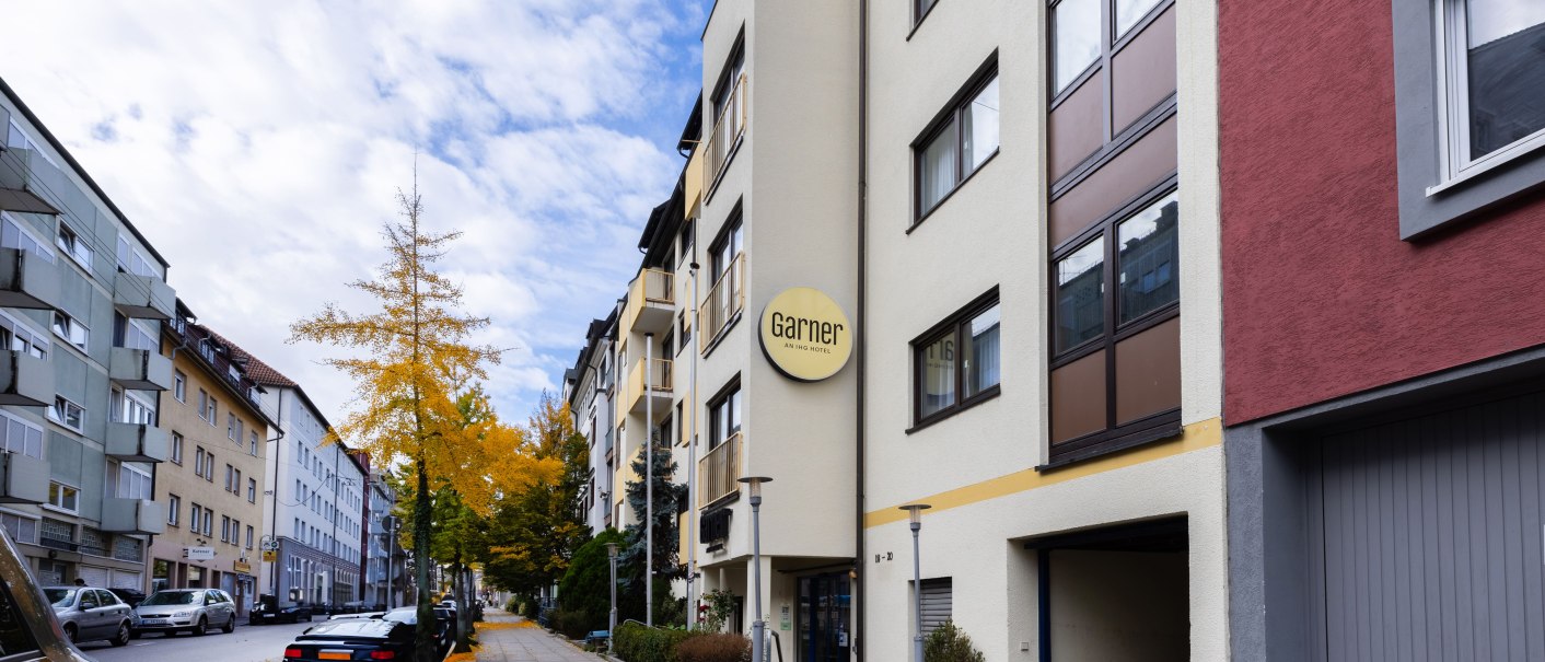 A quiet street lined with multi-story apartment buildings. A tree with yellow autumn leaves stands by the sidewalk. A sign reading "Garner" is visible., &copy; Novum  Hospitality