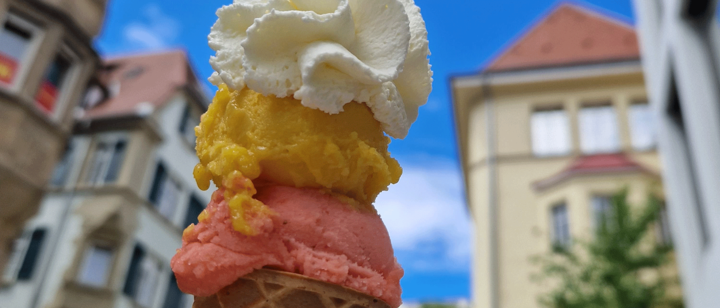 An ice cream cone with three scoops in different colors and whipped cream, against a background of buildings and blue sky., © kesselglueck.de