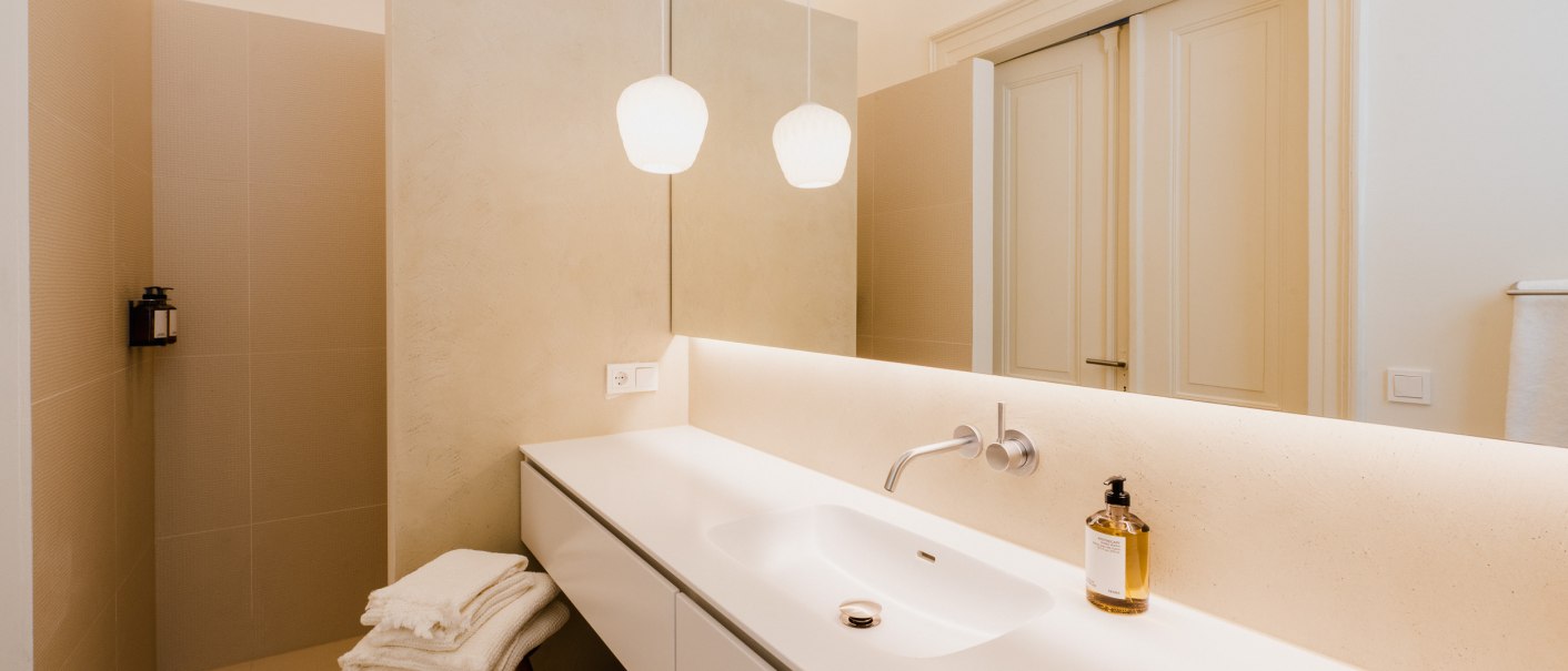 Modernes Badezimmer mit wei&szlig;em Waschbecken, H&auml;ngelampen und Handt&uuml;chern auf einem Hocker. Dezente Beleuchtung und minimalistische Gestaltung., &copy; Peter Oliver Wolff / DAS EDITH