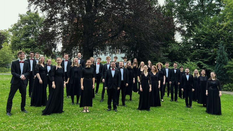 Der Kammerchor der Hochschule f&uuml;r Musik Detmold posiert in eleganter Kleidung auf einer gr&uuml;nen Wiese vor einem gro&szlig;en Baum., &copy; Kammerchor der Hochschule f&uuml;r Musik Detmold