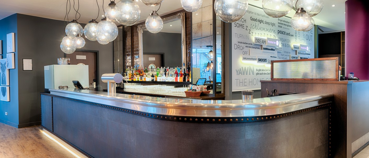 Modern bar with curved counter, hanging glass ball lamps and a shelf full of bottles in the background., © Premier Inn Germany Modern bar with curved counter, hanging glass ball lamps and a shelf full of bottles in the background., © Premier Inn Germany