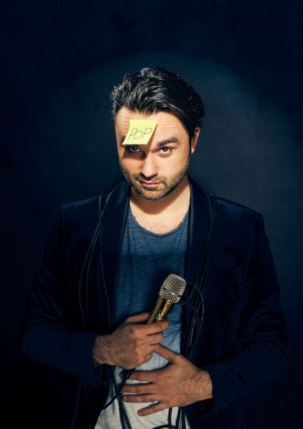 A man is holding a microphone and has a piece of paper with 'POP' written on his forehead. He is wearing a dark jacket and looking directly into the camera., &copy; Theaterhaus Stuttgart e.V.