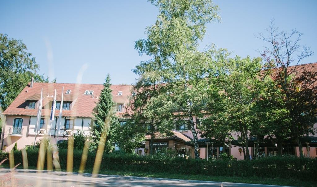 A hotel with a red roof, surrounded by trees, under a blue sky. Flags and a road can be seen in the foreground., &copy; relexa Waldhotel Schatten