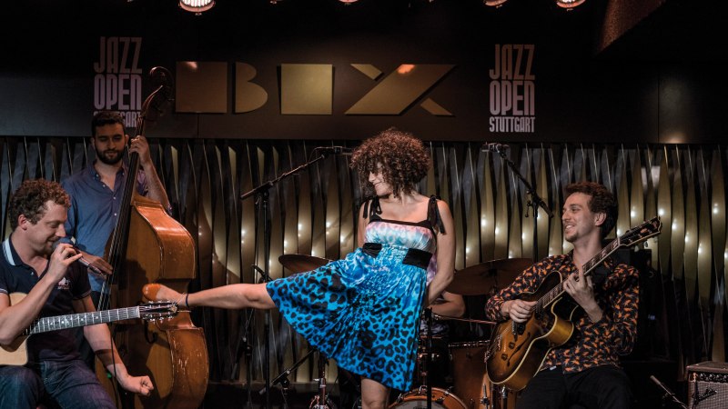 Eine Band spielt im BIX Jazzclub. Ein Gitarrist, ein Kontrabassist und eine Sängerin in blauem Kleid sind zu sehen. "Jazz Open Stuttgart" im Hintergrund., © Peter Steinheisser