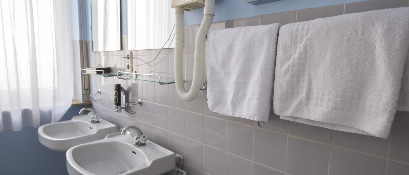 Ein Hotelbadezimmer mit zwei Waschbecken, weißen Handtüchern, einem Haartrockner und einem Spender für Papiertücher an der Wand., © Hotel Ri