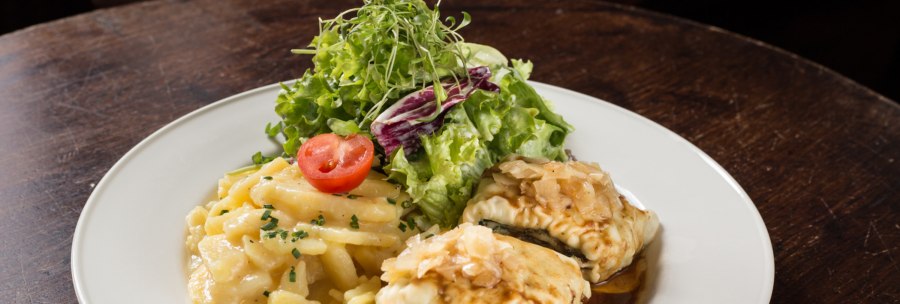 A plate of Maultaschen, potato salad and green salad, garnished with a slice of tomato, on a wooden table., &copy; Amadeus, Stuttgart