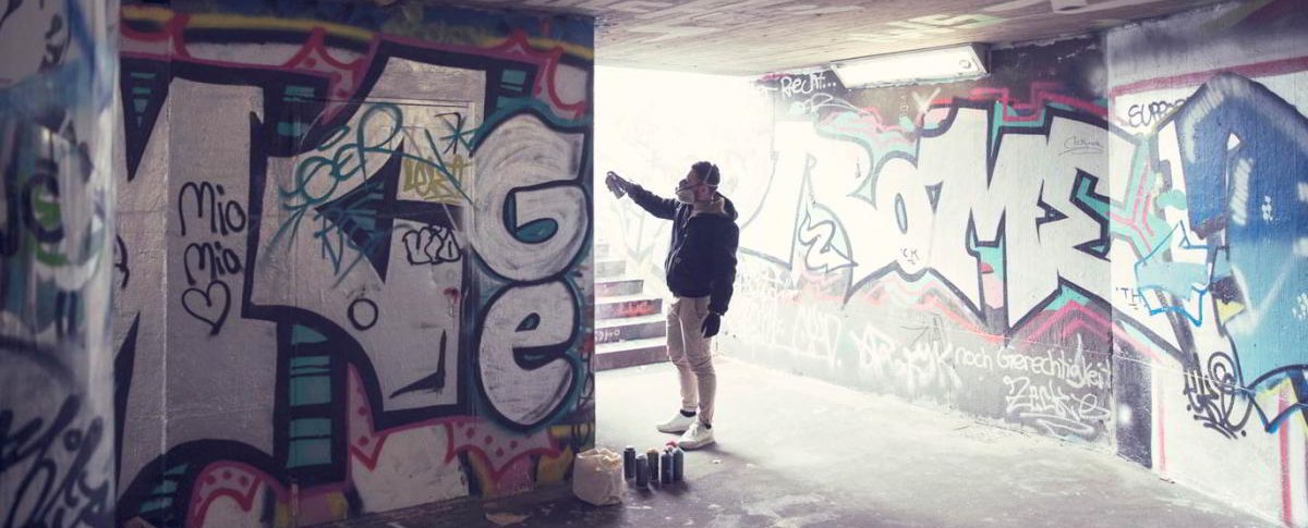 Eine Person sprüht Graffiti in einem Tunnel. Die Wände sind mit bunten Graffitis bedeckt. Auf dem Boden stehen Farbdosen., © hoponopo GmbH
