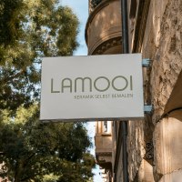 Schild mit 'LAMOOI Keramik selbst bemalen' an einer Steinwand, umgeben von B&auml;umen., &copy; SMG, Sarah Schmid