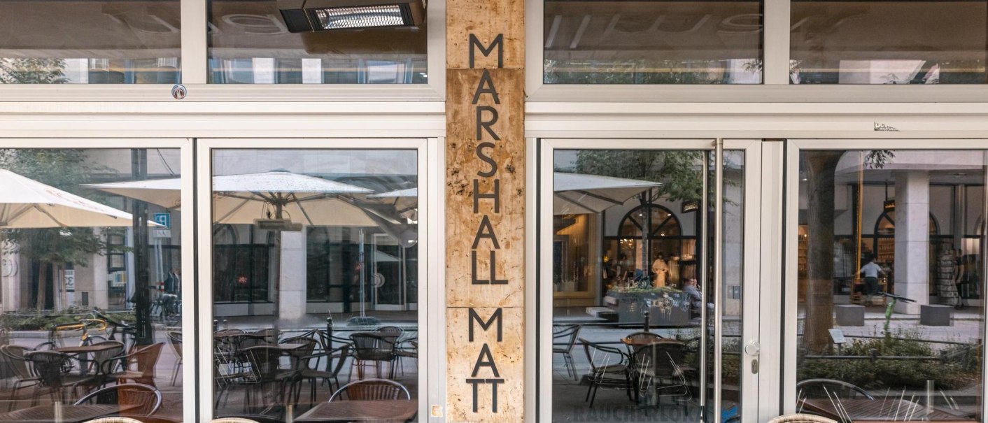 Eingang eines Cafés mit dem Namen Marshall Matt. Glasfront mit Spiegelung von Tischen, Stühlen und Sonnenschirmen im Außenbereich., © Stuttgart Marketing GmbH, Sarah Schmid Eingang eines Cafés mit dem Namen Marshall Matt. Glasfront mit Spiegelung von Tischen, Stühlen und Sonnenschirmen im Außenbereich., © Stuttgart Marketing GmbH, Sarah Schmid