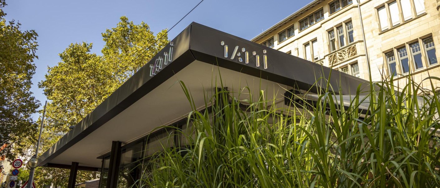 Modern building with the lettering 'Tatti' on the roof. Tall green plants can be seen in the foreground. A historic building in the background., © SMG, Sarah Schmid