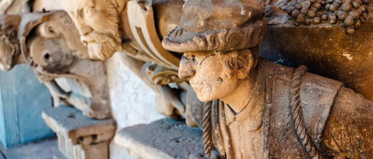 Steinskulptur eines Mannes mit Hut, im Profil. Im Hintergrund sind weitere Skulpturen zu sehen. Die Oberfläche ist verwittert., © Stuttgart Marketing GmbH, Sarah Schmid