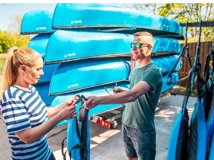 Zwei Personen legen Schwimmwesten an, um sich auf eine Kanufahrt vorzubereiten. Im Hintergrund sind mehrere blaue Kanus gestapelt., &copy; Cool-Tours StattReisen