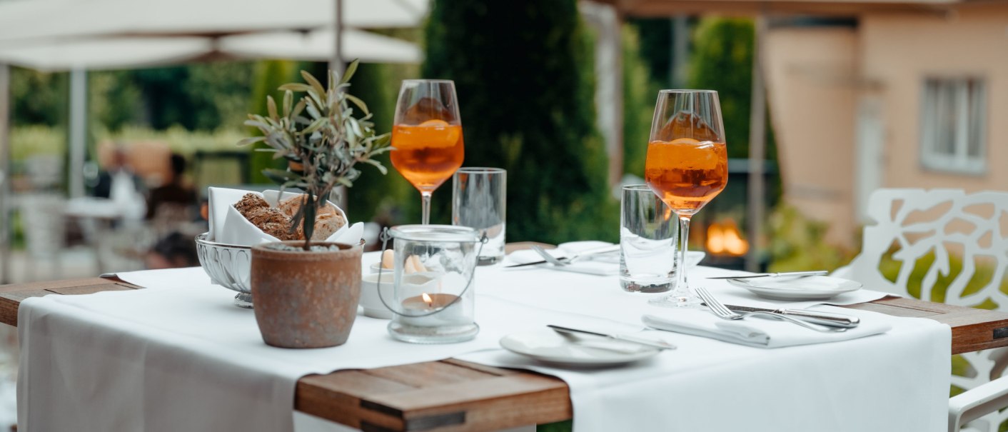 Ein Tisch auf einer Terrasse, gedeckt mit Aperol Spritz, Brot, Kerze und Pflanzen. Im Hintergrund unscharfe Gartenansicht., © Waldhotel Stuttgart GmbH Ein Tisch auf einer Terrasse, gedeckt mit Aperol Spritz, Brot, Kerze und Pflanzen. Im Hintergrund unscharfe Gartenansicht., © Waldhotel Stuttgart GmbH